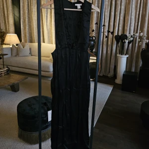 Svart långklänning med volangkant - Elegant svart långklänning från H&M i glansigt material med djup v-ringning och volangkant längs urringningen. Ärmlös modell med markerad midja och dragkedja i ryggen. Perfekt för dig som vill sticka ut med en stilren look.