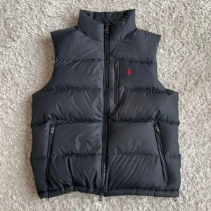 Svart dunväst från Polo Ralph Lauren - Snygg svart dunväst från Polo Ralph Lauren med röd broderad logga på bröstet. Västen har hög krage, tre dragkedjefickor och quiltad design. Perfekt att slänga över en hoodie för en clean look. Västen är perfekt för lager-på-lager nu till hösten.