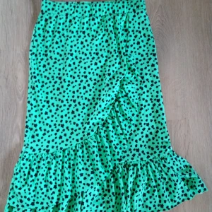 Grön blommig volangkjol - Snygg grön kjol med svarta små blommor och volangkant. Kjolen har omlottkänsla framtill och resår i midjan för skön passform. Perfekt för dig som gillar färg och mönster! Materialet känns lätt och flowy.Kjolen är nästan alldrig andvänd(finns katt i hemmet)💕💕