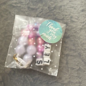 Personligt pärlarmband med namn - Färgglatt armband med pärlor i lila och rosa toner samt vita bokstavspärlor som bildar namnet 'SLEY'. Armbandet har en guldfärgad karbinhake och är perfekt som accessoar till väskan eller nyckelknippan. Levereras i en söt prickig påse.