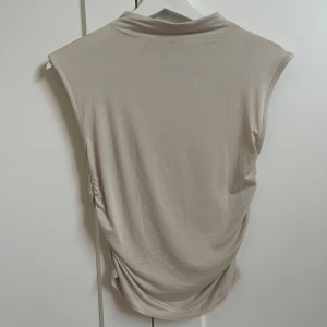Beige topp från Gina Tricot - Säljer en stilren beige  topp från Gina Tricot. Toppen har en mjuk och stretchig känsla med hög hals och enkel design, perfekt att matcha med olika outfits. Den har scrunch i midjan som ger en super snygg figur. Den är aldrig använd så är precis som ny💗
