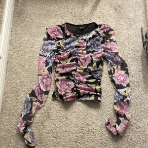 Blommig topp från Gina Tricot - Säljer en blommig topp från Gina Tricot med rosa och lila rosor på en svart bakgrund. Toppen har långa ärmar och en rynkad detalj framtill som ger en fin passform. Perfekt för att liva upp garderoben med lite färg och mönster!