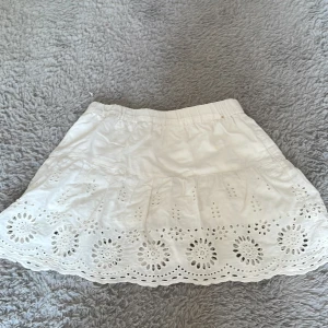Vit kjol med broderade detaljer från Kappahl - Supersöt vit kjol från Kappahl med elastisk midja och fina broderade blommönster längs nederkanten. Kjolen är kort och har en luftig känsla, perfekt för varma dagar.