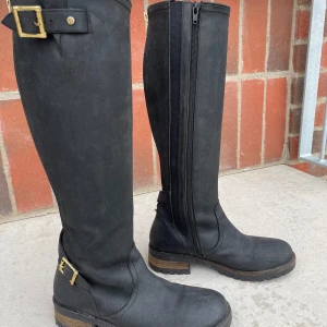 Svarta höga stövlar - Ett par svarta höga stövlar/boots från Dasia. Storlek 40 och skaft W2 (ca 37 cm). Grov sula, guldfärgade justerbara dragkedjor och spännen upptill och vid ankeln. Inköpta runt 2015 för ca 1500kr. Sparsamt använda och i fint skick, har stått oanvända i flera år.