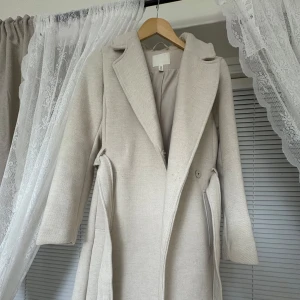 Beige kappa med klassisk krage - Säljer en stilren beige kappa med klassisk krage och knappar i midjan. Jackan har långa ärmar och två fickor framtill. Perfekt för dig som vill ha en tidlös och elegant look. Använd två gånger från H&M.