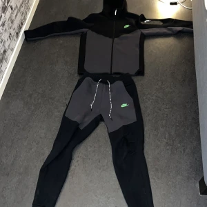 Svarta och mörkgrå Nike tracksuit  - Snygg nike tracksuit från jd sport i svart och mörkgrått med grönt Nike-märke Perfekta för en avslappnad stil.  använd 4 gånger ungefär,köpte den från jd sport för ungefär 3 månader sen. Den har bara legat i min garderob sen dess. Kvitto finns kvar!! 