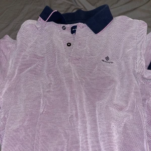 Ljuslila pikétröja från GANT - Säljer en ljuslila pikétröja från GANT med mörkblå krage och rosa detalj. Tröjan har korta ärmar, knappar framtill och är tillverkad i 100% bomull. Klassisk och stilren modell med diskret logga på bröstet. Helt äkta.