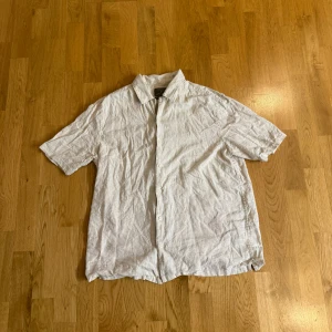 Vit linneskjorta med kort ärm - Det är en luftig skjorta tillverkad av linne, ett naturligt material som andas bra och håller dig sval under varma dagar. Färgen är klassiskt vit, vilket ger ett fräscht och tidlöst intryck. Den korta ärmen gör skjortan ledig men ändå uppklädd, perfekt för både strandpromenader och sommarfester. Skjortan kan ha knappar framtill och kanske en bröstficka som detalj. 