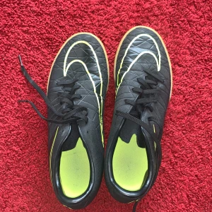 Svarta inomhus fotbollsskor med neongula detaljer från Nike - Säljer ett par svarta inomhus fotbollsskor från Nike med neongula detaljer och snörning. Skorna har en klassisk rund tå och är designade för bra grepp på planen. Perfekta för dig som vill sticka ut lite extra på matchen!