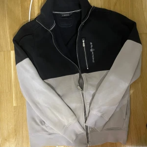 Svart och grå ziptröja från Sail Racing - Tvåfärgad ziptröja från Sail Racing i svart och ljusgrått. Jackan har hög krage, dragkedja framtill och en bröstficka med dragkedja. Logga på bröstet och ribbade muddar vid ärmslut och nederkant.