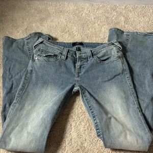 Blå jeansbyxor från True Religion - Säljer ett par klassiska blå jeans från True Religion med fem fickor och snygga kontrastsömmar. Byxorna har en ikonisk ficka med lock och knapp bak samt en ljus tvätt för en avslappnad look.