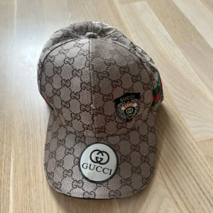 Beige keps från Gucci - Säljer en beige keps från Gucci med klassiskt GG-monogram över hela kepsen. Kepsen har gröna och röda ränder på sidan, svart läderrem bak och tydliga Gucci-loggor framtill. VÄLDIGT LIKT ORGINAL 