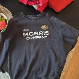 Mörkblå tröja från Morris - Säljer en snygg mörkblå tröja från Morris med broderad logga och text på framsidan. Tröjan har långa ärmar och ribbade kanter vid hals, ärmslut och nederkant. Perfekt för en stilren look.