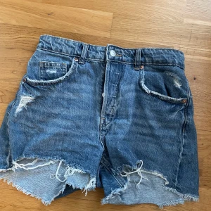 Blå jeansshorts från H&M - Säljer ett par coola blå jeansshorts från H&M med slitna detaljer och fransiga kanter. Perfekta för en avslappnad stil. De har en klassisk femficksdesign och knappgylf.
