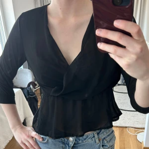 Svart omlottblus med trekvartsärm - Säljer en svart omlottblus med trekvartsärm. Blusen har v-ringning och är tillverkad i ett tunt, lätt och något transparent material. Sitter snyggt i midjan och har en feminin passform. Perfekt att styla med jeans eller kjol. Aldrig använd. Kan bäras i S och XS. 