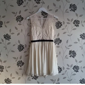 Vit spetsklänning med svart bälte - Elegant vit klänning med spetsdetaljer och ett svart band i midjan. Klänningen är ärmlös och har en v-ringad framdel. Perfekt för sommarens festligheter. Asos storlek 38.