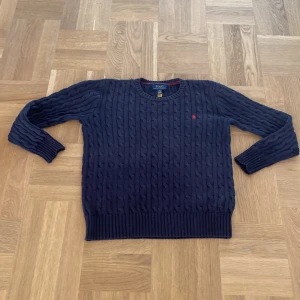 Mörkblå kabelstickad tröja från Polo Ralph Lauren - Säljer en mörkblå kabelstickad tröja från Polo Ralph Lauren med rund halsringning och röd broderad logga på bröstet. Tröjan har långa ärmar och ribbade muddar vid ärmslut och nederkant. Perfekt för en klassisk och stilren look.