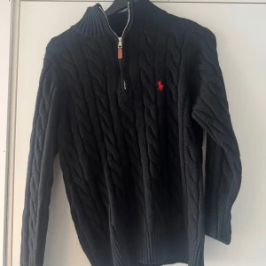 Svart kabelstickad tröja från Ralph Lauren - Säljer en svart kabelstickad tröja från Ralph Lauren med halv dragkedja och hög krage. Tröjan har lång ärm och röd broderad logga på bröstet. Perfekt för kyligare dagar och snygg till jeans eller chinos.