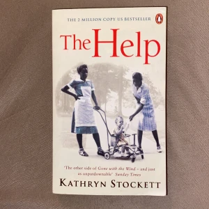 The Help - Säljer romanen 'The Help' av Kathryn Stockett. Boken är på engelska och har ett vitt omslag med röd text och ett svartvitt foto på två kvinnor och ett barn. Pocketutgåva från Penguin Books. Perfekt för dig som gillar gripande berättelser om vänskap och samhälle.