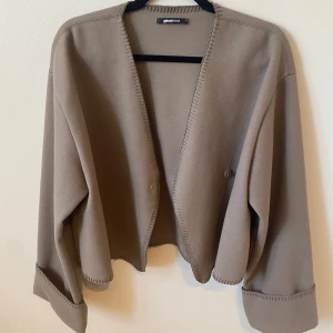 Beige cardigan jacka från Gina Tricot - Säljer en beige cardigan jacka från Gina Tricot. Jackan har en öppen design utan krage, med dekorativa sömmar längs kanterna och vida ärmar. Perfekt för lager-på-lager och enkel att styla till olika outfits.