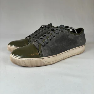 Grå och olivgröna sneakers från Lanvin - Säljer ett par stilrena sneakers från Lanvin i grå mocka med olivgröna detaljer i blankt material på tån och hälen. Skorna har snörning och en vit sula som ger en snygg kontrast. Perfekta för dig som gillar exklusiva och unika sneakers.