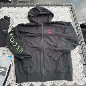 Svart hoodie med dragkedja från Nike - Säljer en svart hoodie från Nike med dragkedja och huva. Den har en röd Nike-logga på bröstet och texten 'SWOOSH' i grönt på ärmen. Perfekt för en avslappnad stil.