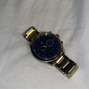 Guldfärgad klocka med blå urtavla - Snygg guldfärgad klocka från Tommy Hilfiger med en stilren blå urtavla. Klockan har en metallarmband och flera små urtavlor för extra funktionalitet. Perfekt för den som vill ha en elegant accessoar. Batteriet är dock slut  till salu 