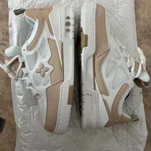 Vita och beige sneakers från Louis Vuitton - Snygga vita och beige sneakers från Louis Vuitton med ikoniska detaljer och logotyp. Skorna har en rund tå och snörning, vilket ger en klassisk look. Perfekta för den stilmedvetna som vill ha något extra i garderoben. Det går att diskutera om priset.