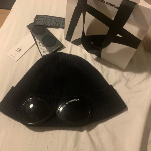 Cp company mössa - Snygg svart mössa från C.P. Company med unika glasögondetaljer framtill. Den är ribbad och ger en cool och modern look. Perfekt för att sticka ut i mängden och hålla sig varm samtidigt. Väldigt bra skick, köpt på nk och allt stämmer bra med den. Är öppen för prisförslag det är bara att höra av sig. Allt og medföljer och är såklart äkta