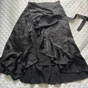 Svart omlottkjol från Chelsea - Elegant svart omlottkjol från Chelsea med volangdetaljer. Kjolen har ett knytband i midjan och är tillverkad i ett mjukt, glansigt material som ger en lyxig känsla. Perfekt för att skapa en stilren look. Aldrig använd🦋🥰