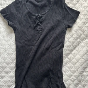 Svart ribbad topp med knyt - Säljer en svart ribbad topp med korta ärmar och snygg knytning framtill. Perfekt för en stilren look. Passar till både jeans och kjol!