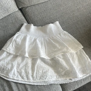 Vit kjol  - Superfin och somrig kjol från SRN HLT med vackra broderade blommor och volangdetaljer. Kjolen har en elastisk midja för bekväm passform och är perfekt för varma dagar. Den broderade designen ger en romantisk touch. Storlek 146/152 men passar som en Xs/S