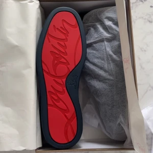 Mörkblå sneakers från Christian Louboutin - (Pris kan diskuteras)          Säljer ett par mörkblå sneakers från Christian Louboutin med klassisk röd sula och ovandel i mocka. Skorna har rund tå, platt sula och snörning. Kommer med originalpåse och låda.