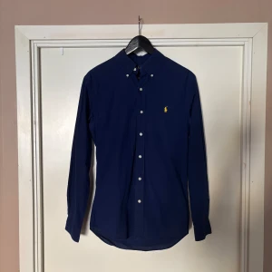 Mörkblå skjorta från Ralph Lauren - Säljer en klassisk mörkblå skjorta från Ralph Lauren med knappar framtill och broderad gul logga på bröstet. Skjortan har lång ärm och button-down krage, perfekt för en stilren look.