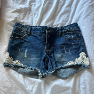 Lågmidjade jeansshorts  - Snygga blå jeansshorts med fransig kant och vita spetsdetaljer på sidorna. Klassisk femficksmodell med knapp och dragkedja framtill. Perfekta nu till sommaren🫶🏻