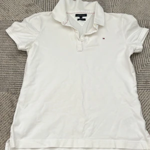 Vit pikétröja från Tommy Hilfiger - Säljer en klassisk vit pikétröja från Tommy Hilfiger med korta ärmar och knappslå. Tröjan har en liten logotyp på bröstet och en stilren krage. Perfekt för en avslappnad och stilren look.