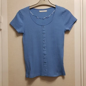 Blå ribbad topp med låsas knappar strl S - Säljer en blå ribbad topp med korta ärmar och låsas knappar framtill. Tillverkad i 95% bomull och 5% elastan för en bekväm passform. Perfekt för en avslappnad stil.