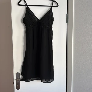 Svart klänning från BlendShe - Snygg svart klänning från BlendShe med smala axelband och v-ringning. Klänningen har dekorativa pärlor under bysten och ett lätt, flödande tyg som ger en elegant look. Perfekt för en kväll ute eller en speciell tillställning.