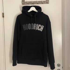 Svart hoodie från Hoodrich med tryck - Svart hoodie från Hoodrich i storlek S med stor logga i vitt och grått framtill. Tröjan har huva med snörning, magficka och mjukt fleecefoder på insidan. Perfekt för dig som gillar streetwear och vill ha en skön och stilren look.
