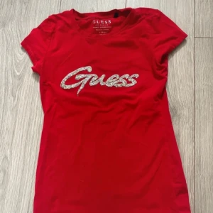 Röd t-shirt med glitterlogo från Guess - Snygg röd t-shirt från Guess med korta ärmar och glittrigt Guess-tryck på bröstet. Modellen är figurnära och har rund hals. Toppenskick och sitter verkligen jättesnyggt på! Meddela gärna om du har några frågor🥰