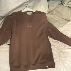 Brun sweatshirt från Les Deux - Säljer en stilren brun sweatshirt från Les Deux med broderad logga på bröstet. Tröjan har rund halsringning, långa ärmar och ribbade muddar. Perfekt för dig som gillar en clean och enkel look.