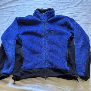 Blå teddyjacka från Haglöfs - Blå teddyjacka från Haglöfs med svarta detaljer på sidor och ärmar. Jackan har hel dragkedja framtill, hög krage och är tillverkad i mjuk fleece. Perfekt för kyliga dagar och outdoor-häng. justerbar nederkant. Pris kan diskuteras 