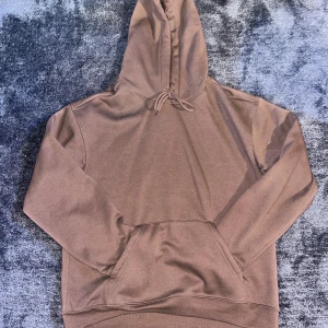 Brun hoodie med huva och ficka - Säljer en enkel brun hoodie med huva och snörning. Tröjan har en stor magficka framtill och är långärmad med ribbade muddar. Perfekt för chill dagar och passar till jeans eller joggers. Materialet känns mjukt och skönt mot huden.