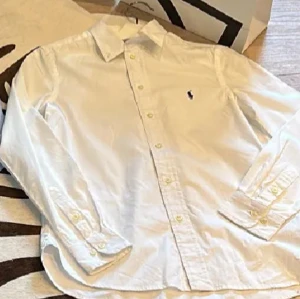 Vit skjorta Polo Ralph Lauren slim fit - Vit långärmad skjorta från Polo Ralph Lauren i slim fit-modell. Klassisk krage, knappar framtill och broderad svart logga på bröstet. Tillverkad i mjuk bomull som känns skön mot huden. Perfekt för dig som gillar stilrena och tidlösa plagg.