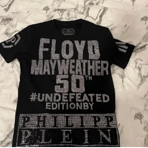 Philipp Plein Mayweather t-shirt svart - Svart t-shirt från Philipp Plein med stora silvriga strasstexten 'Floyd Mayweather 50th #Undefeated Edition' fram och bak. Kortärmad med rund hals och exklusiv design. Materialet är mjuk bomull och detaljerna är glittriga, vilket ger en lyxig vibe.