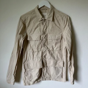 Beige overshirt från A Day's March - Stilren beige overshirt från A Day's March i 100% bomull. Jackan har fyra stora fickor framtill, klassisk krage och knäppning med knappar. Perfekt lager-på-lager-plagg med avslappnad vibe och snygg passform. 1F9
