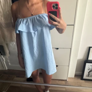 Ljusblå offshoulder kortklänning - Supersöt ljusblå kortklänning med offshoulder och volang upptill. Klänningen har en avslappnad passform och är perfekt för varma dagar. Materialet känns lätt och luftigt, och den offshoulder-stilen ger en trendig vibe. Passar dig som gillar enkel men snygg stil.