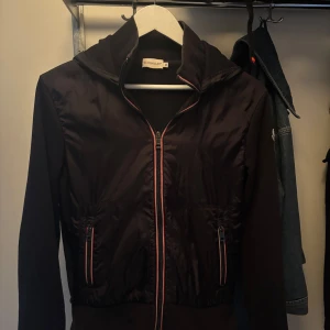 Svart Moncler Windbreaker - Svart Windbreaker från Moncler med huva och dragkedja framtill. Jackan har två fickor med dragkedja och Moncler-logga på ärmen. Materialmix med glansig polyester framtill och mjukare tyg på ärmar och rygg. Perfekt för dig som gillar stilren och sportig look. 