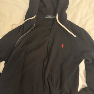 Svart hoodie från Polo Ralph Lauren - Svart hoodie från Polo Ralph Lauren med dragkedja, vit snörning i huvan och röd broderad logga på bröstet. Klassisk passform och mjukt material, perfekt för chill dagar. Fickor framtill för extra bekvämlighet.