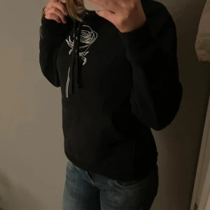 Svart hoodie med ros från The Cool Elephant - Svart hoodie från The Cool Elephant med vit broderad ros på bröstet. Tröjan har huva med snörning och en klassisk känguruficka framtill. Mjuk insida och långärmad modell💕
